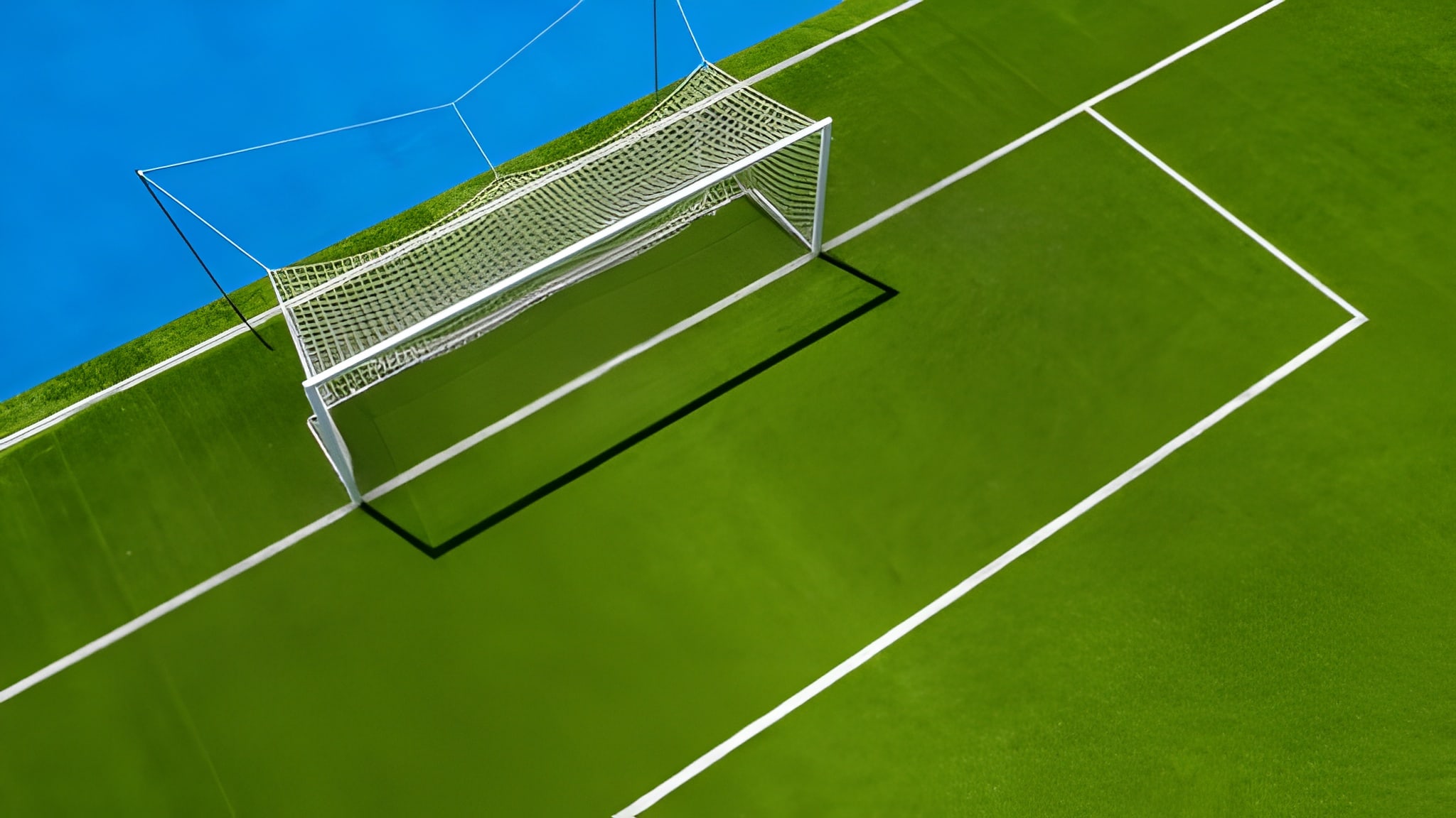 An overhead, high-angle view of a white soccer goal netting on a vibrant green field with crisp white boundary lines, set against a bright blue track border.