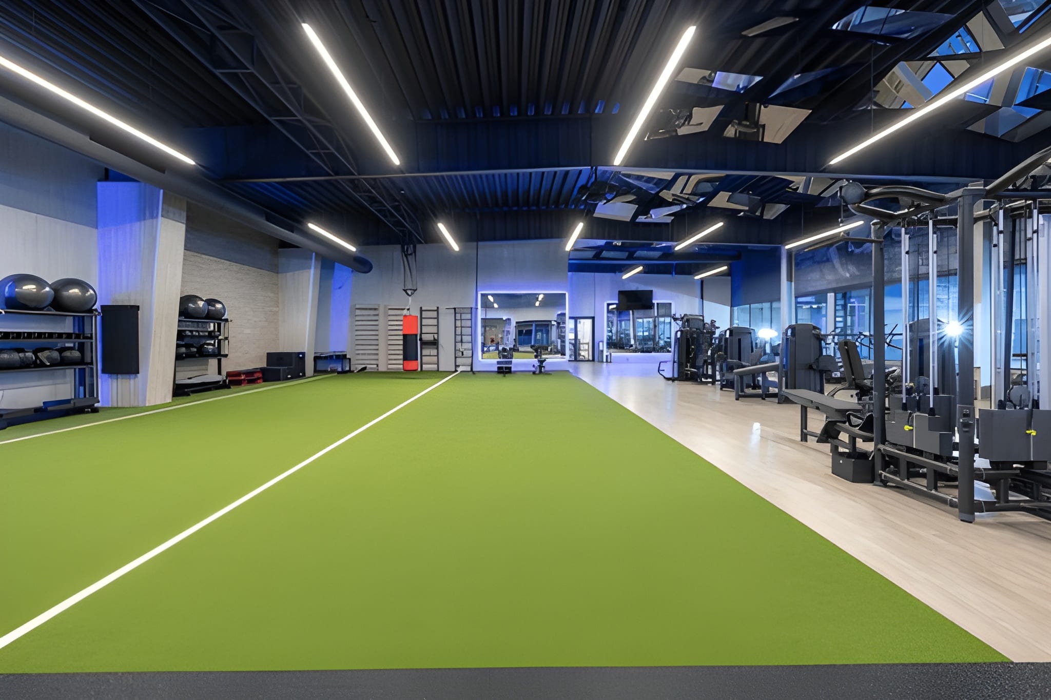 An indoor fitness center featuring a long, green artificial turf agility track with a white line, flanked by exercise equipment and modern overhead lighting.