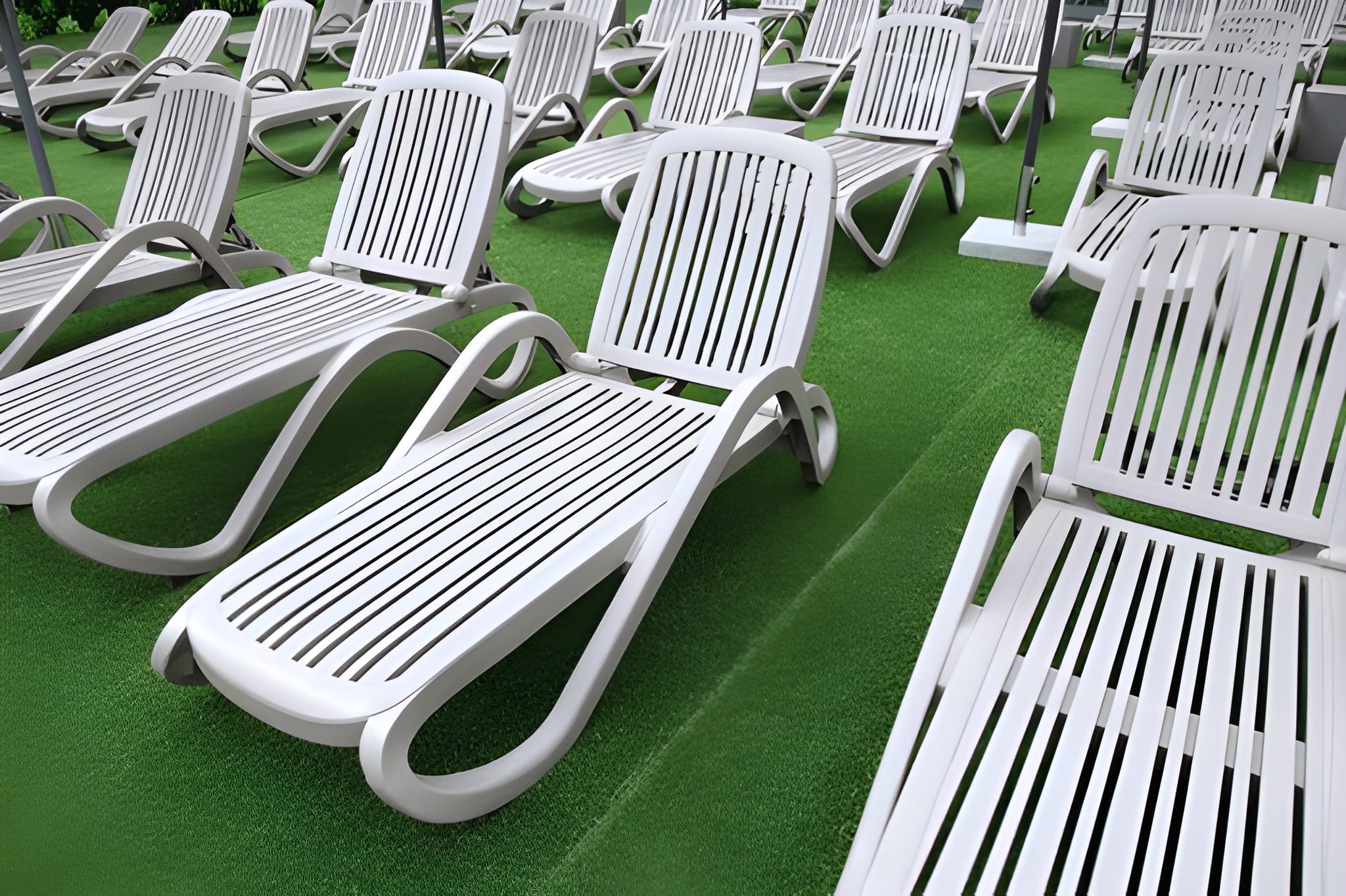 Several rows of white plastic slatted lounge chairs arranged neatly on top of a bright green artificial grass surface.