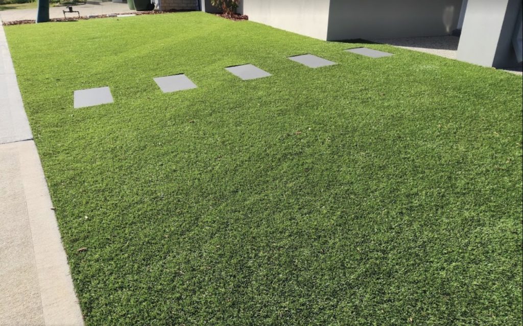 Modern residential front yard featuring vibrant artificial grass integrated with a row of square grey stepping stones next to a concrete driveway