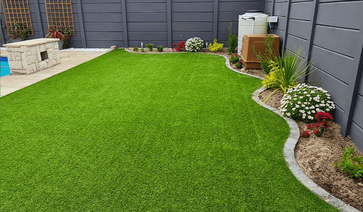 Clean artificial grass lawn in a backyard featuring a curved stone border, small garden bed, and a modern dark grey fence