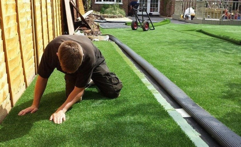 Landscaping installer on his knees smoothing out and fitting synthetic turf tightly against a wooden privacy fence during an outdoor residential installation project