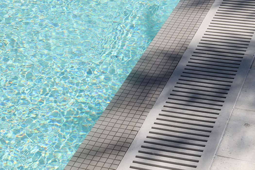 A slotted trench drain system installed along the tiled edge of a swimming pool to manage water overflow and drainage.