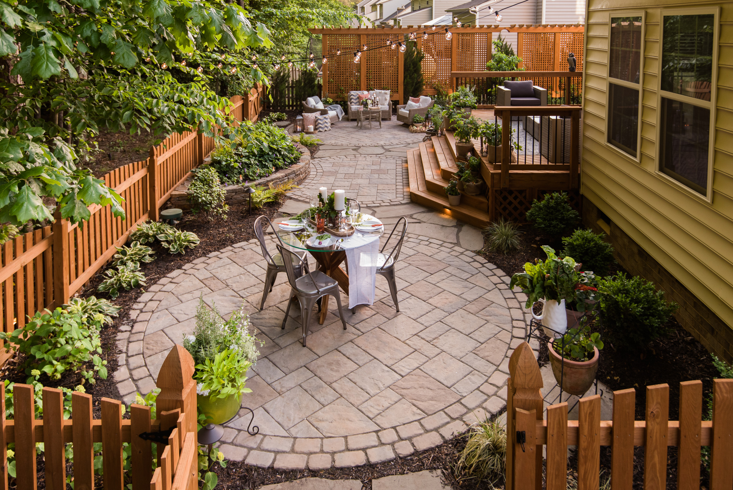 A circular stone paver patio in a fenced backyard with a small dining table, connected to a wooden deck with seating.